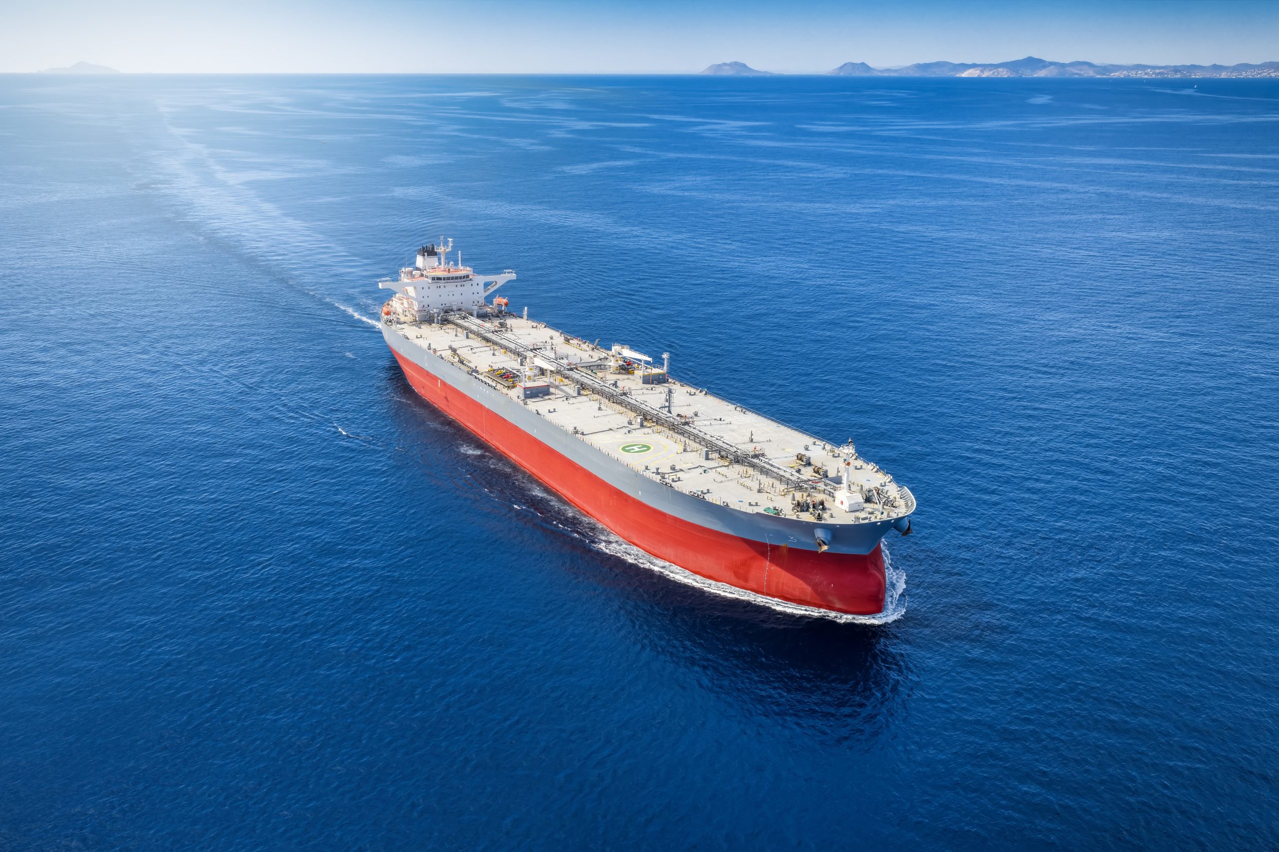 A large red and white tanker sailing through calm blue ocean with distant landmasses on the horizon.