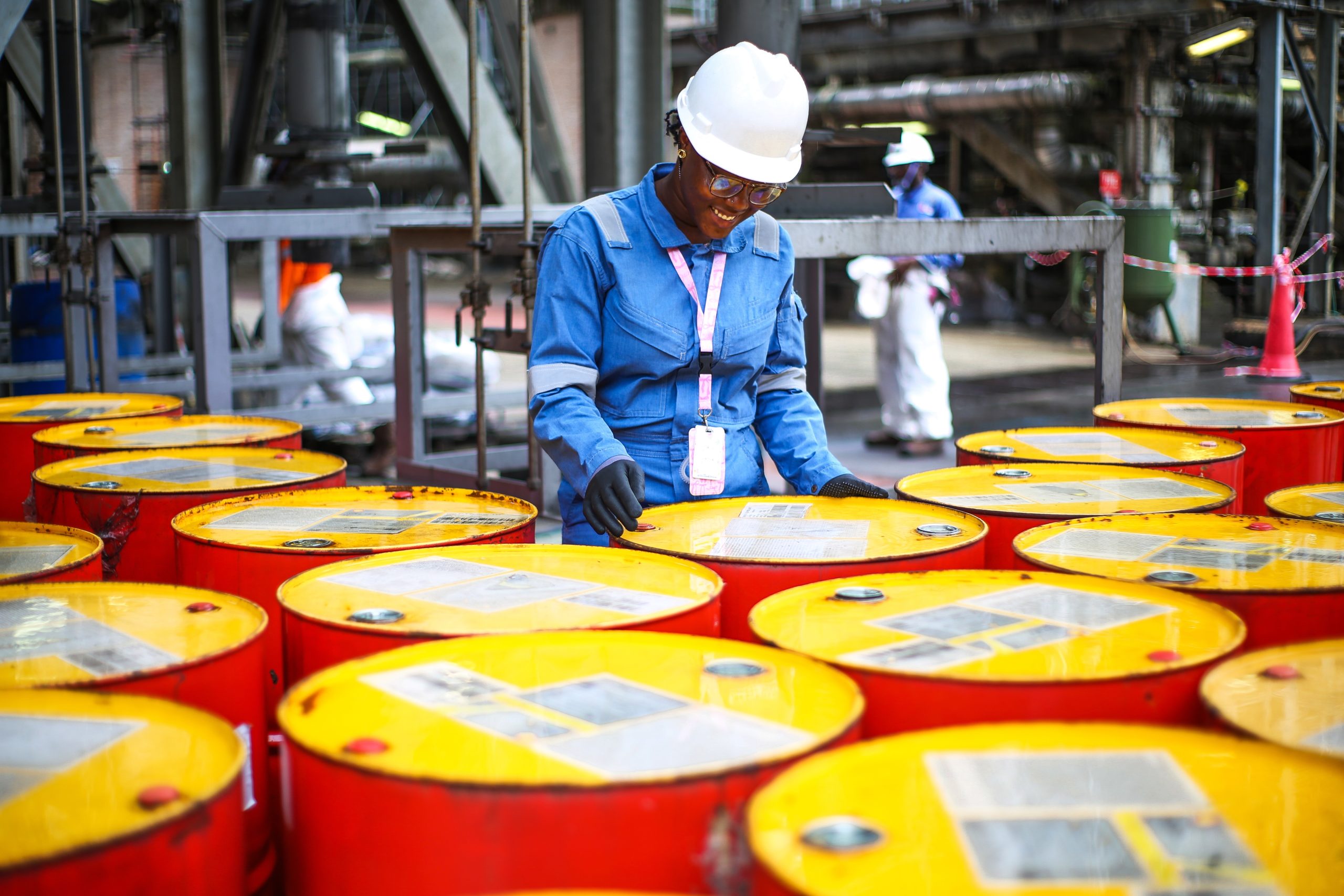 Female worker in blue coveralls and a white hard hat inspects yellow drums in an industrial plant, safety glasses on.